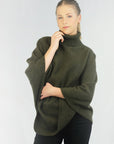 A woman wearing a green turtleneck Merinosilk rib neck plain poncho, paired with black pants.