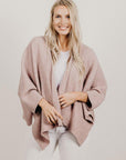 Woman wearing a pink poncho over a white top and pants on a plain background