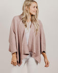 Woman wearing a pink knitted cardigan over a white top and pants on a plain background