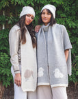 Two women wearing beanies and scarves with animal patterns standing in front of a green leafy background