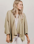 Woman wearing a beige knitted cardigan over a white top and pants on a plain background