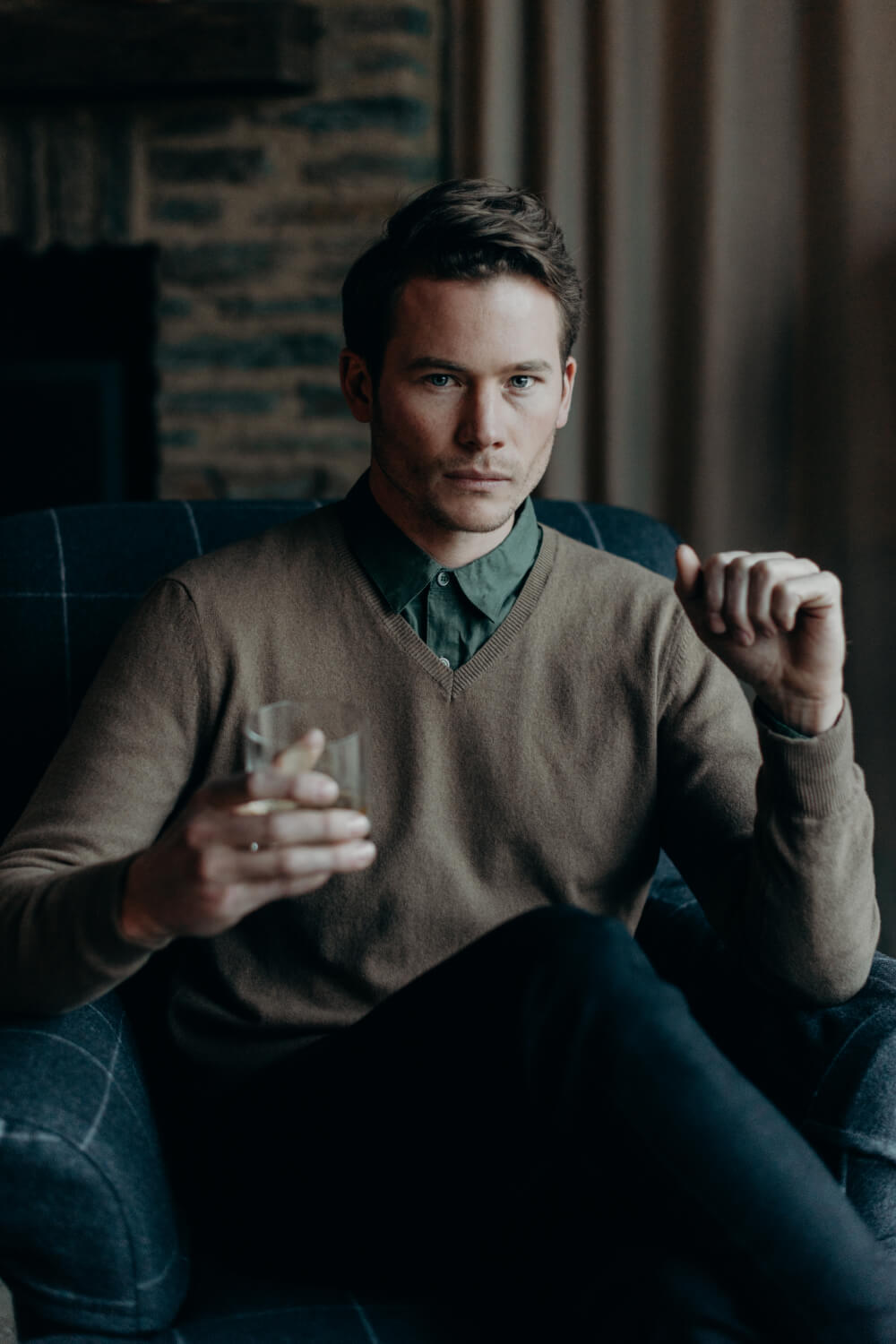 Man sitting in a chair holding a glass, wearing a brown sweater and dark jeans, with a blurred background.