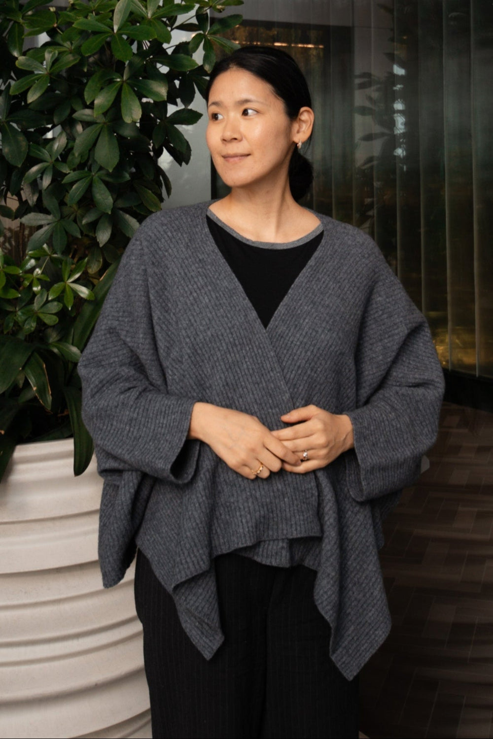 Woman wearing a gray poncho standing next to a plant indoors