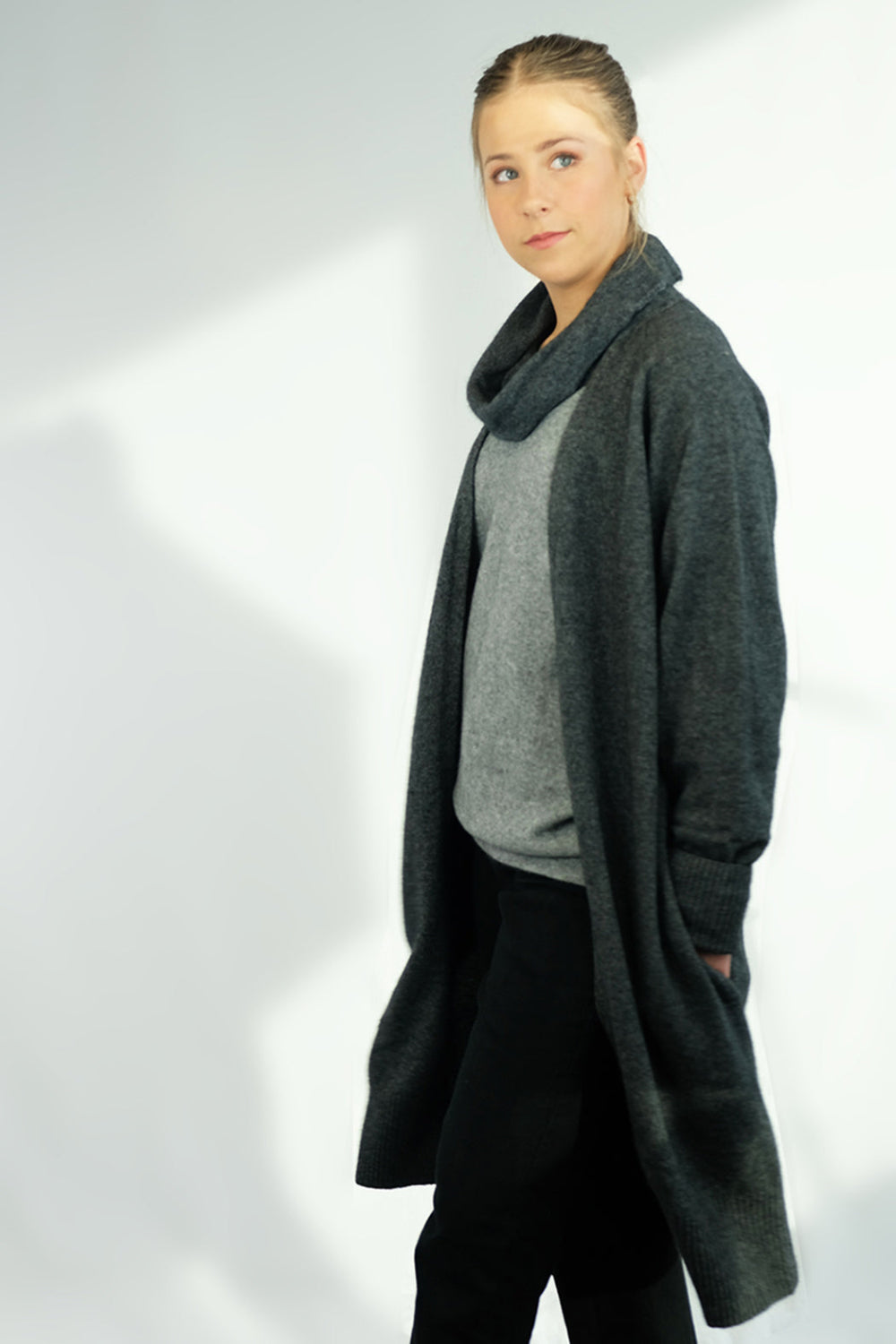 A woman standing, wearing a long gray cardigan with hidden side pockets over a gray sweater and black trousers.