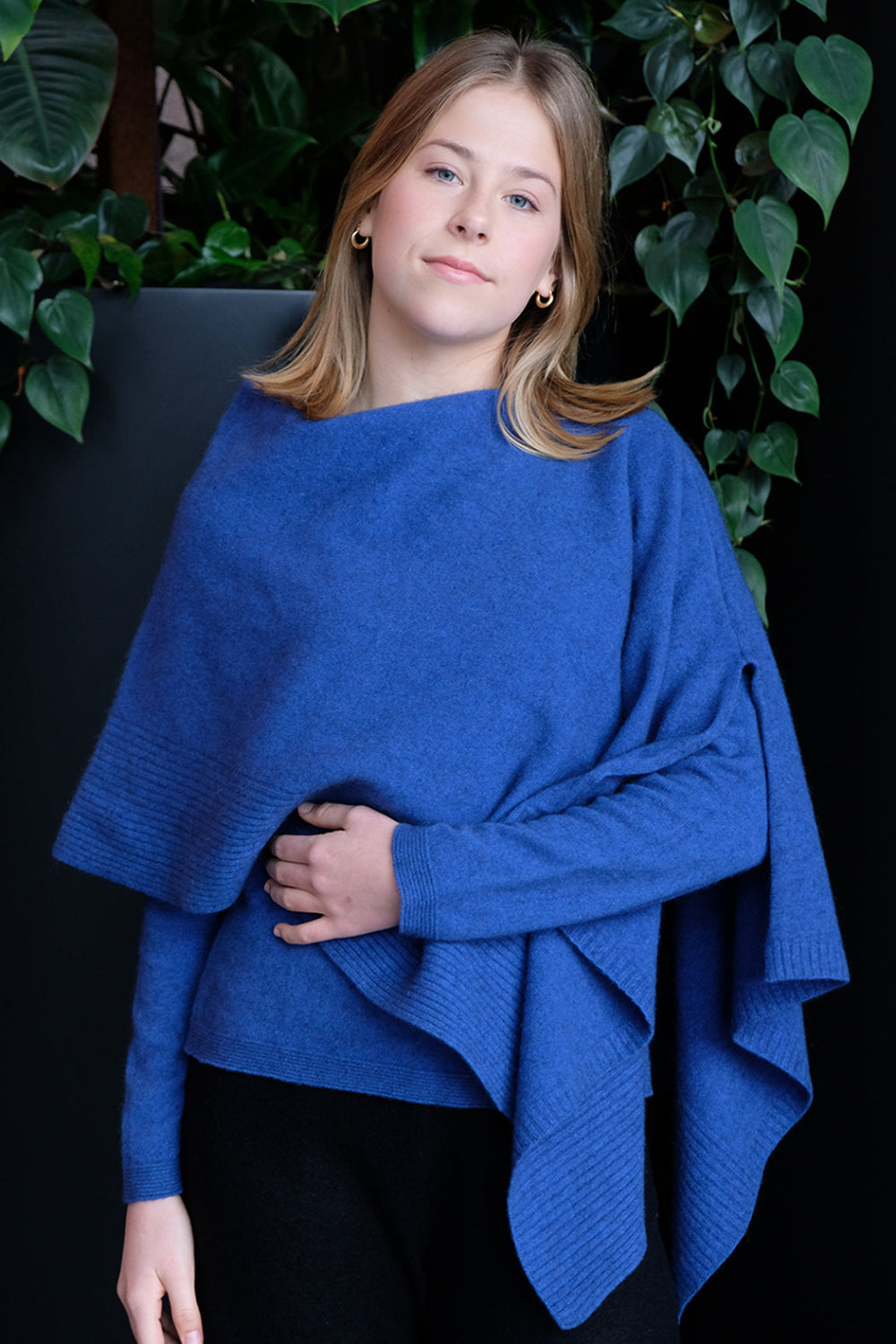 Woman wearing a blue poncho against a dark background with greenery