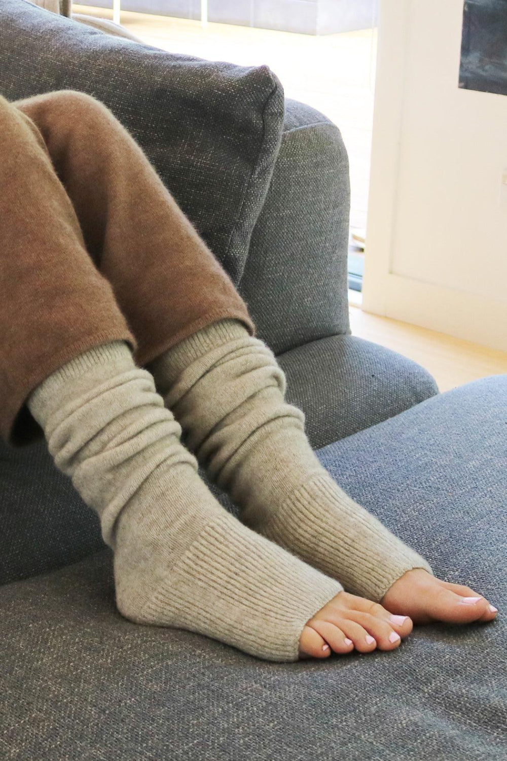 A pair of beige leg warmers worn on a person's legs, resting on a grey sofa.
