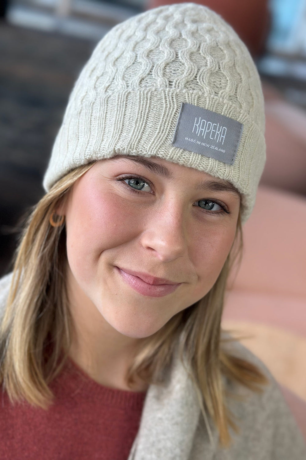 A person wearing a beige cable knit beanie with the &#39;Kapeka&#39; brand label visible on the front.