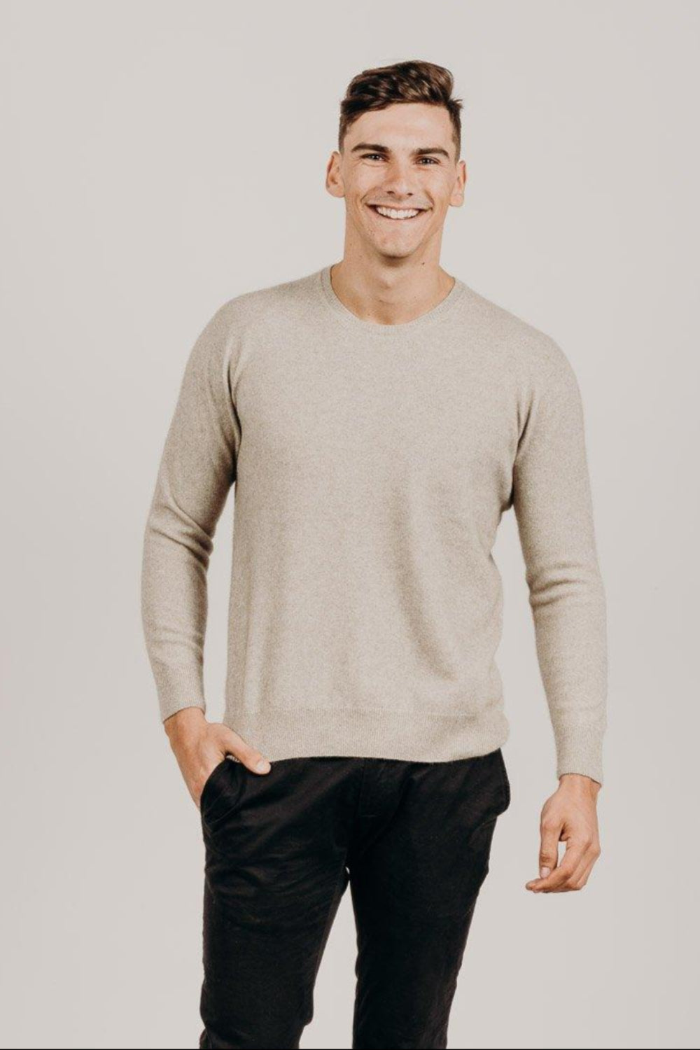 Man wearing a beige sweater and black pants against a plain background