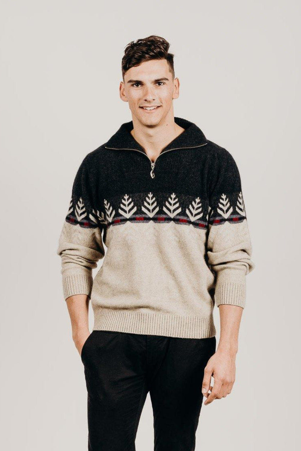 A man wearing a Merinosilk Silver Fern zip Sweater with a half-zip front, paired with black trousers. The sweater has a silver fern pattern on a beige background.