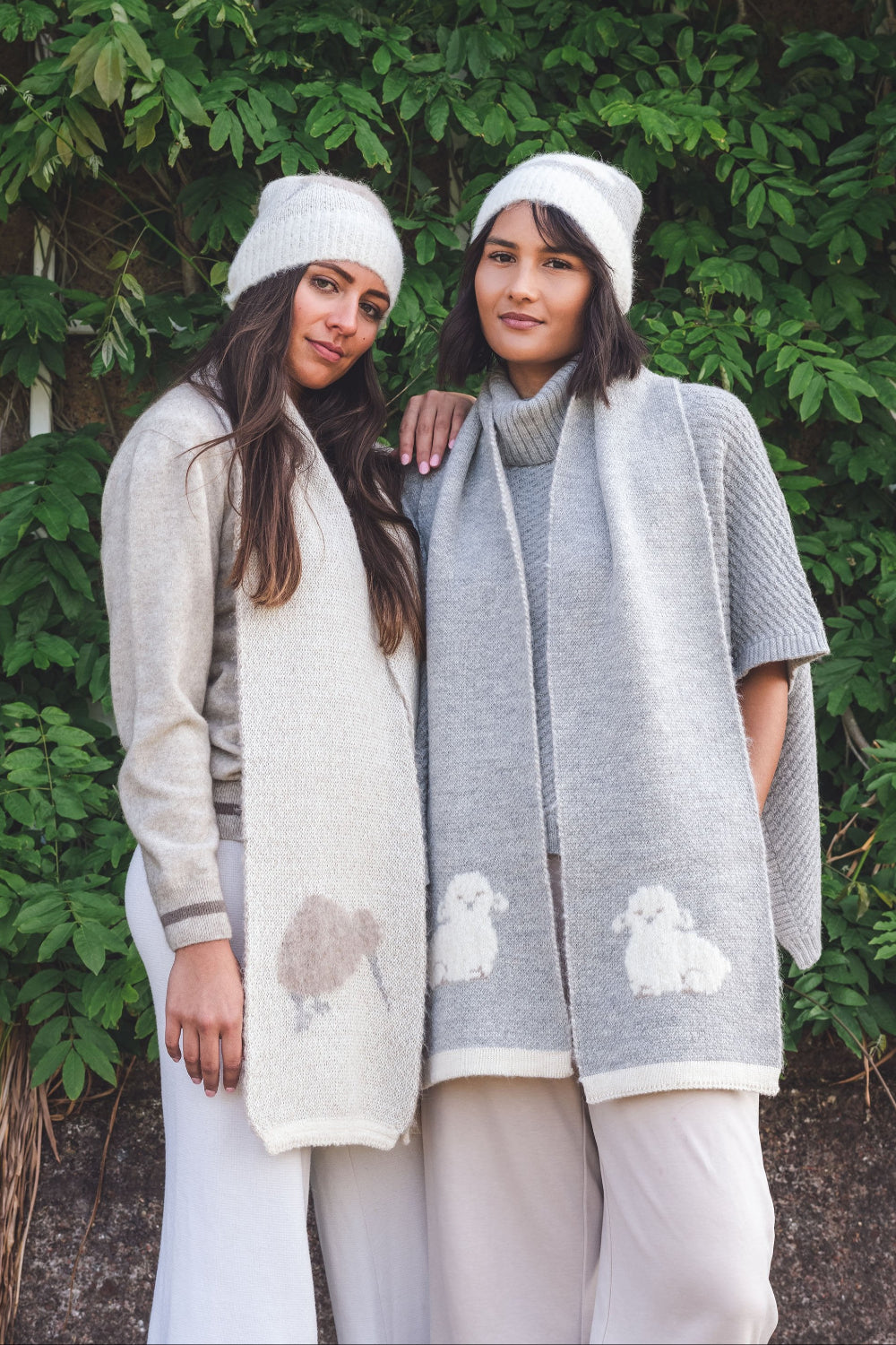 Two women wearing beanies and scarves with animal patterns standing in front of a green leafy background