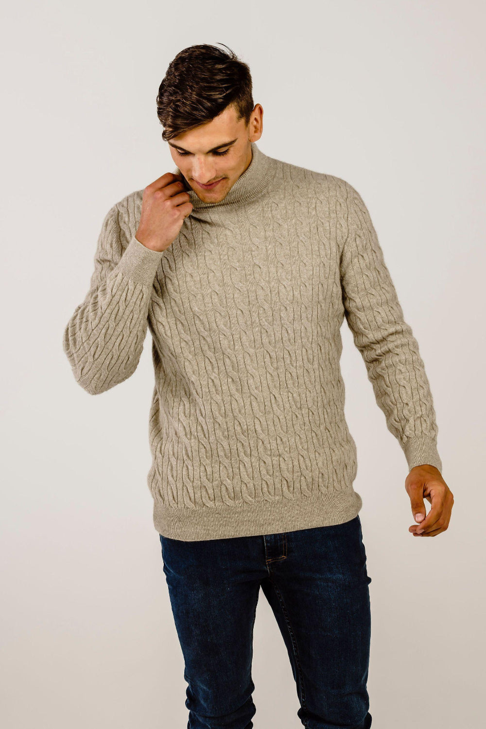 Man wearing a beige cable knit sweater and dark jeans on a plain background