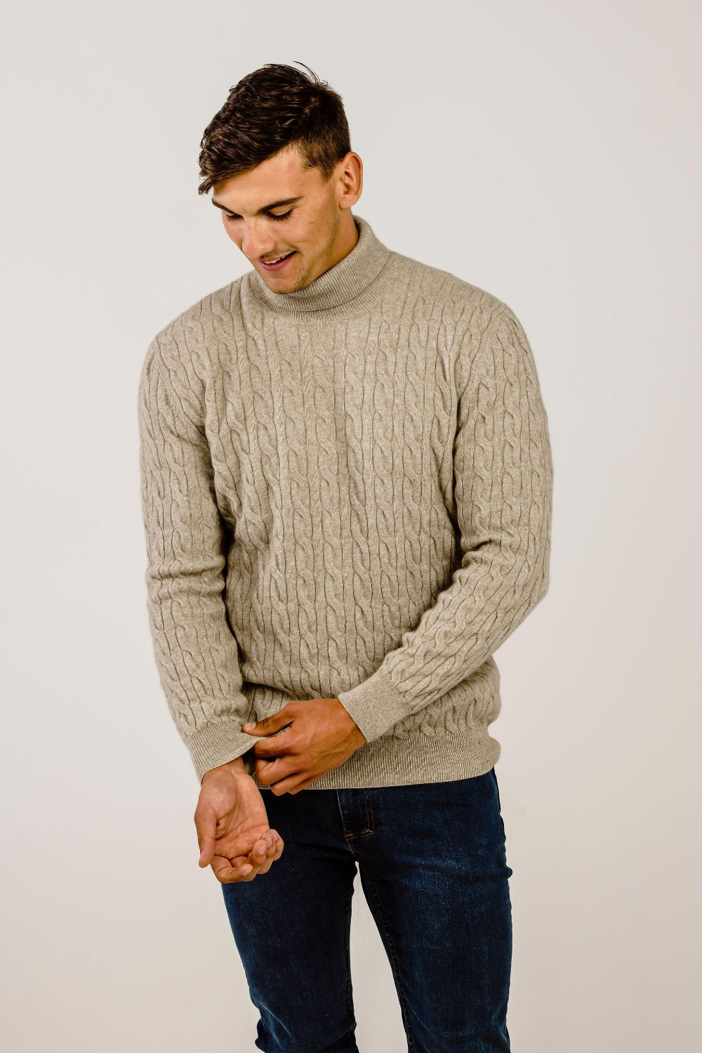 Man wearing a beige cable knit sweater and dark jeans against a plain background