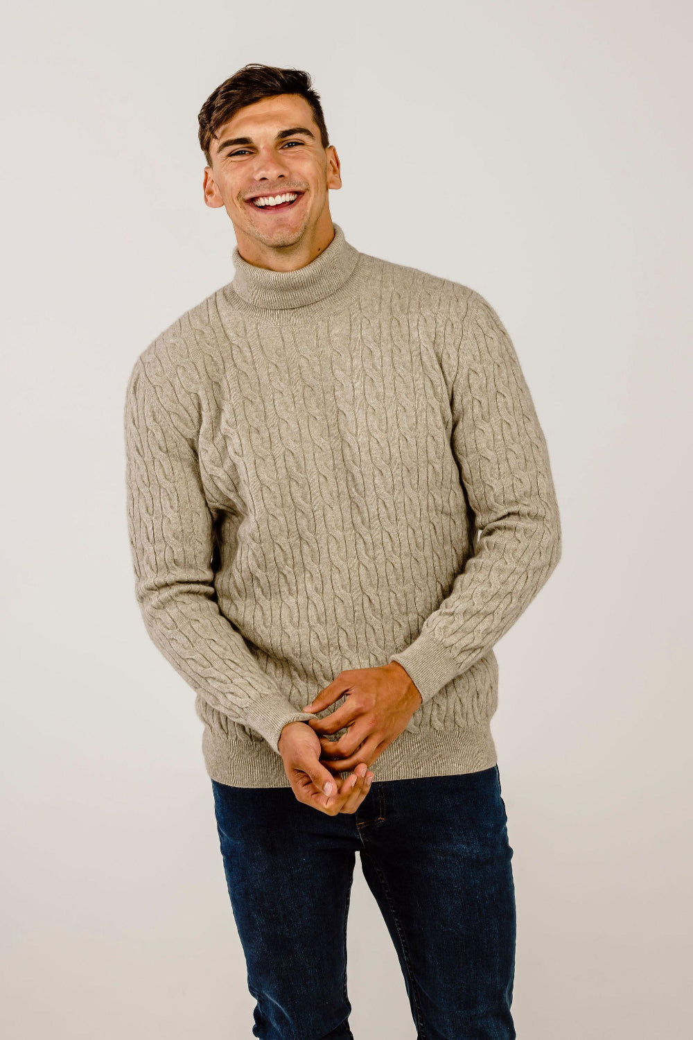 Man wearing a beige cable knit sweater and dark jeans against a plain background
