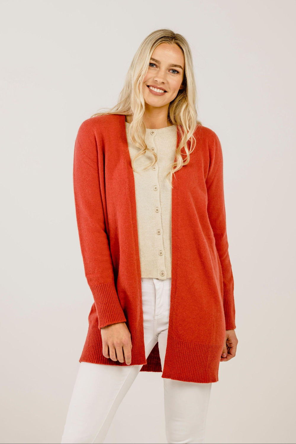 Woman wearing a red cardigan over a beige top and white pants on a light background