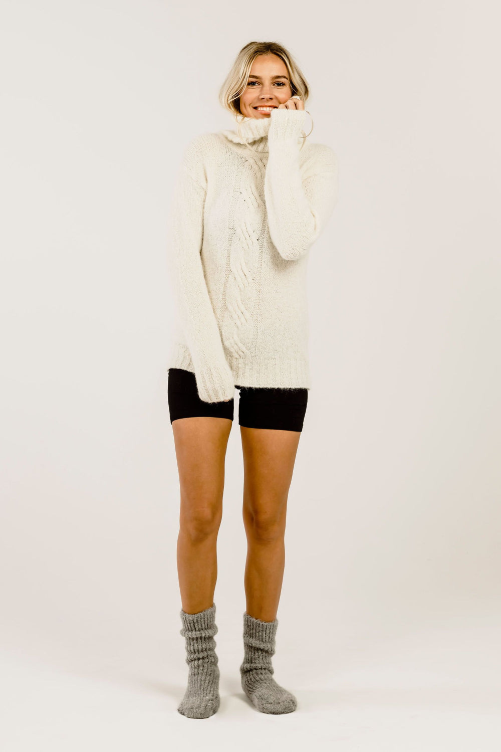 Woman wearing a cream cable knit sweater over a black pants with gray socks on a white background