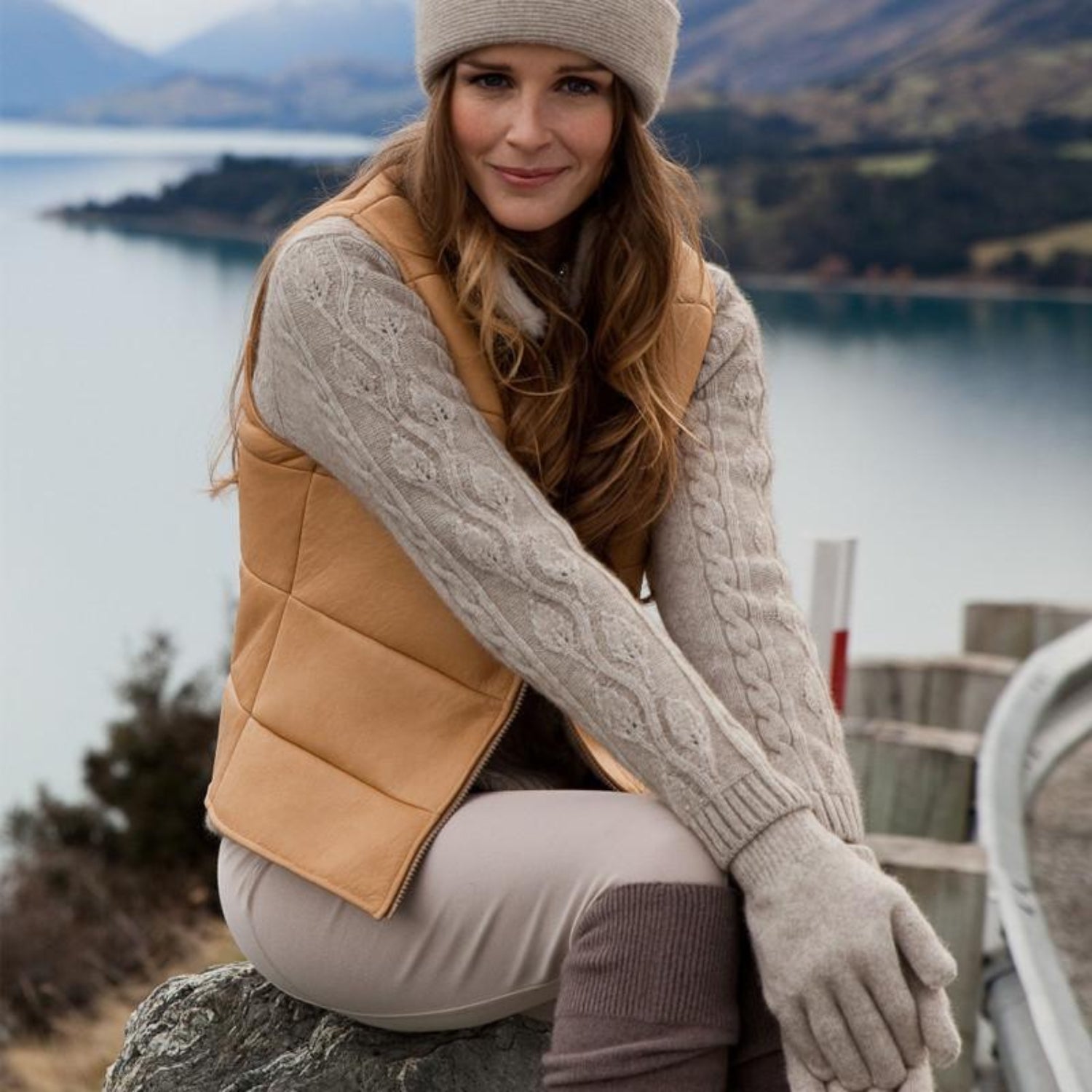 Woman in a beige vest and gloves sitting by a lake with mountains in the background